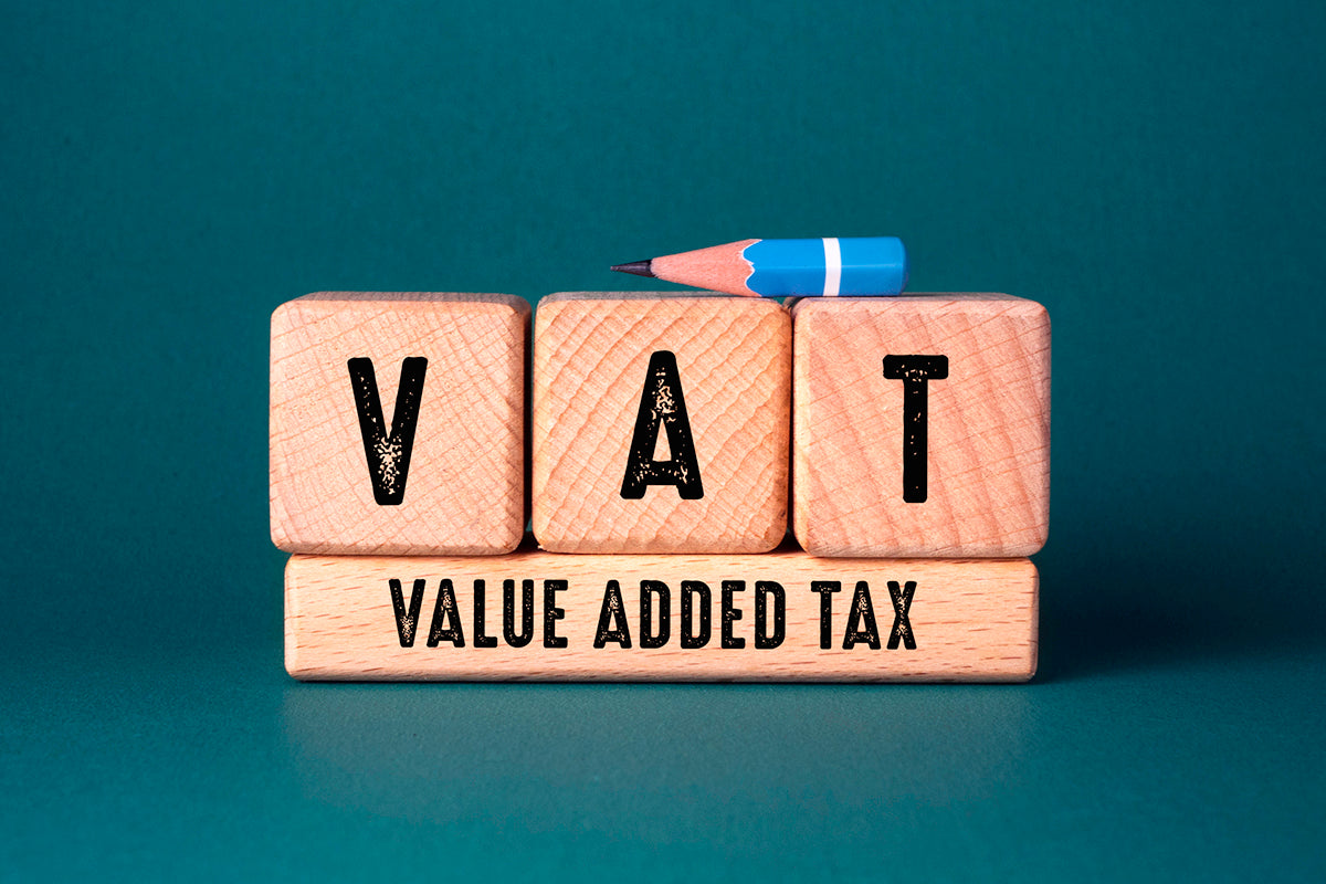 Three square wooden blocks with the letters V A T printed on them, sitting on a thin rectangular block with 'value added tax' printed on it. A tiny stub of pencil rests on top of the blocks.