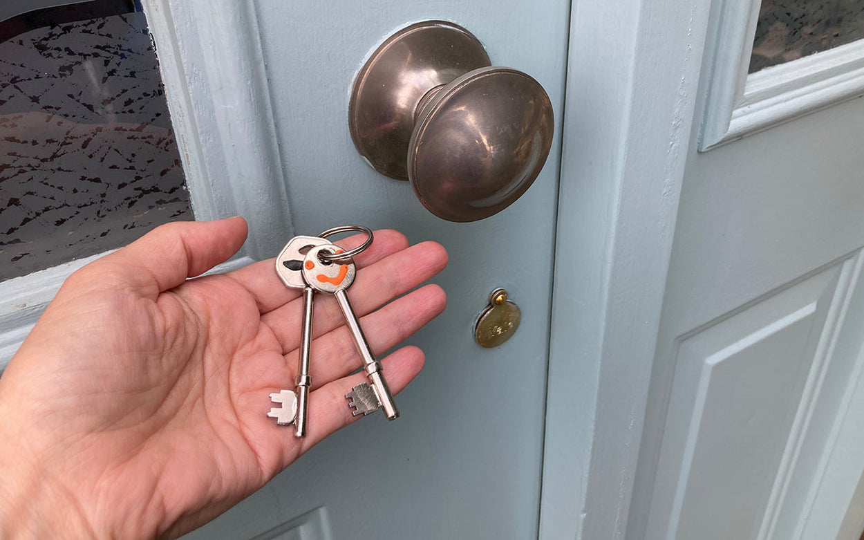 A person's hand holding two similarly shaped door keys in front of a pale blue exterior door. One key has a horizontal line of black tacti-mark on it, the other has a smiley face marked in orange tacti mark