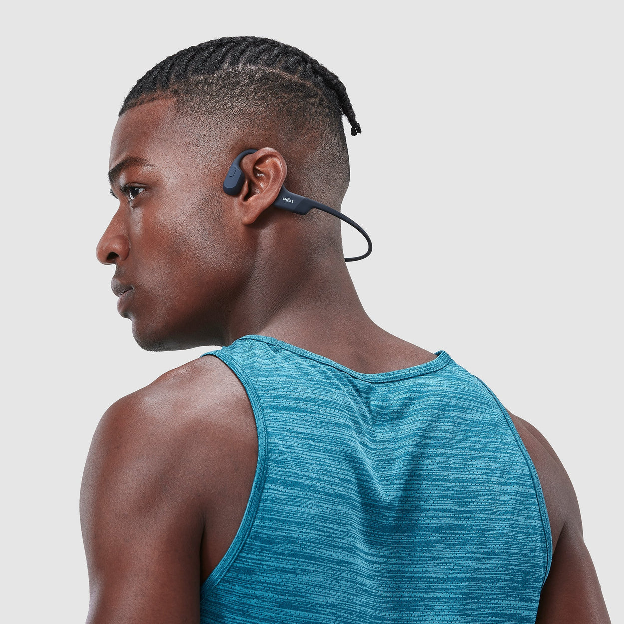 A young black man in a turquoise sports vest stands with his back to the camera and his head turned to show his side profile. The OpenMove headphones sit around the back of his head with the bone-conduction speaker resting in front of his ear