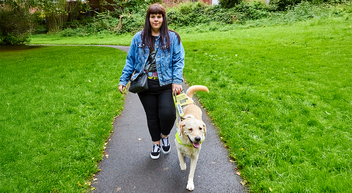 Lorna walking with her guide dog. She has brown hair, is wearing a denim jacket, black jeans, and black trainers.