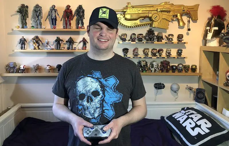 Sightless Kombat, a young white man in a dark t-shirt with a skull and blue gear wheel on it and baseball cap, holds a games console controller in front of shelves containing models of characters from games and science fiction franchises.