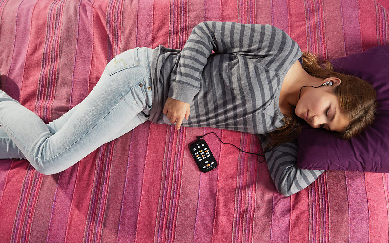 A teenage girl is lying on a bed with a pink cover, while listening to something on a DAISY player