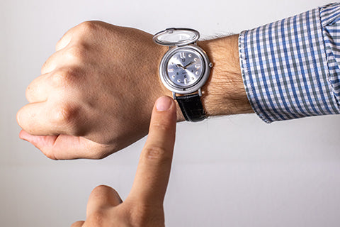 A person holding out their arm to show a tactile watch on their wrist. The case is open and the person's other finger is outstretched to read the time using the tactile markings on the watch face.