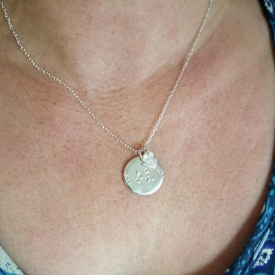 Disc-shaped silver necklace with ‘love’ in braille and charm being worn