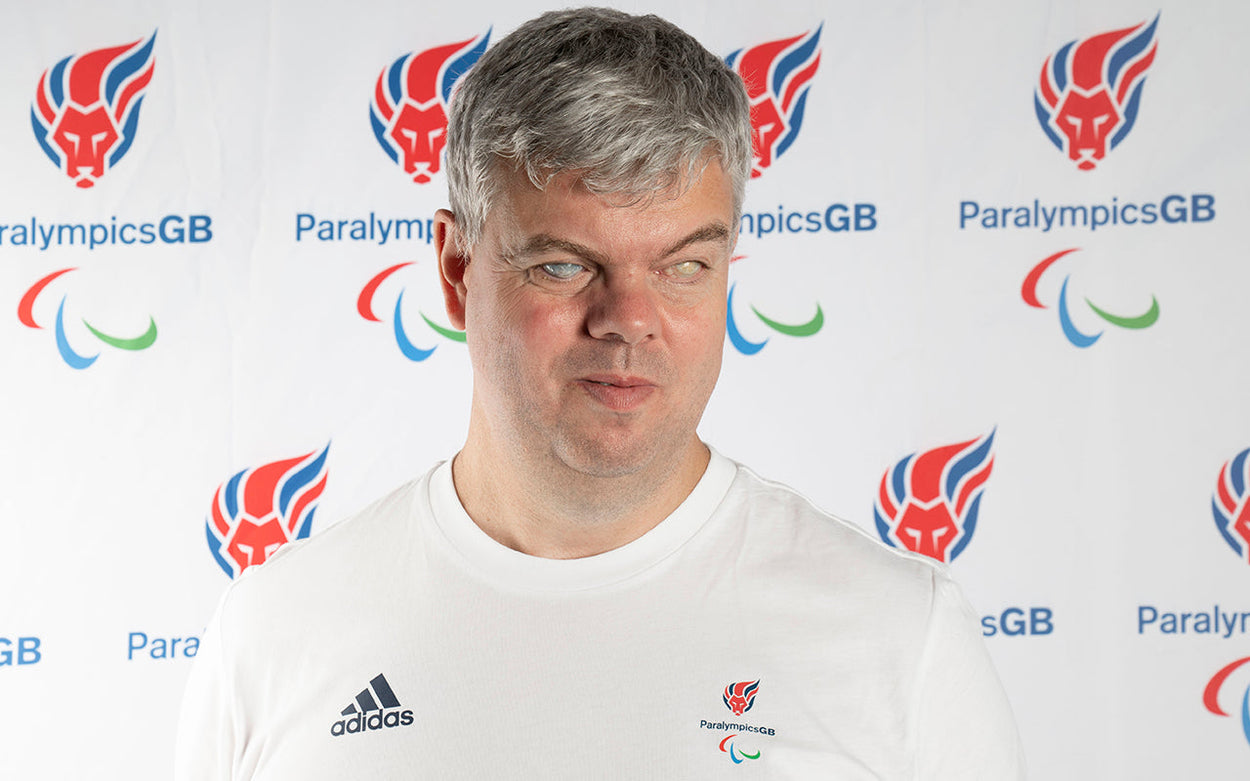 David Clarke in a white ParalympicsGB top in front of a white backdrop with repeating Paralympics GB logo (a red, white and blue lion's head with a mane that resembles flames)