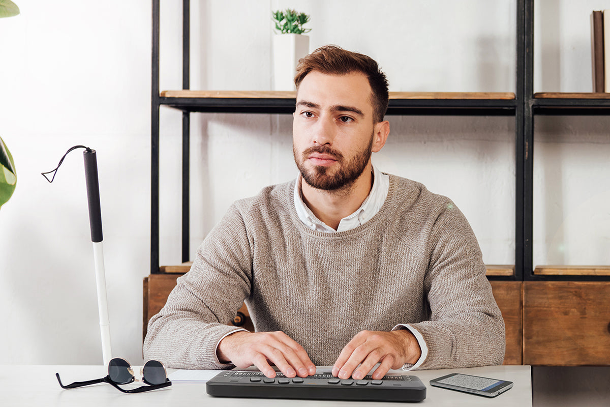 A young white man with dark hair and short beard sitting at a white desk using a braille notetaker