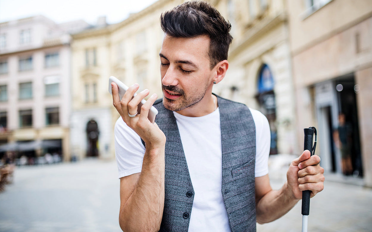 A man standing on a street using an iPhone in one hand while holding a white cane in the other. 