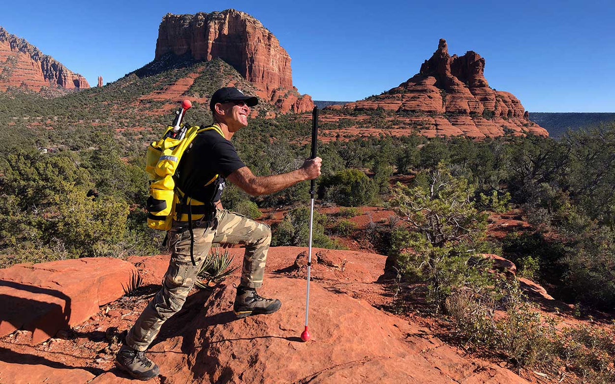 A deeply tanned man smiles while stepping up on to a rock on an Arizona trail, supported by an all terrain cane. Behind him are red sandstone buttes and a deep blue sky.
