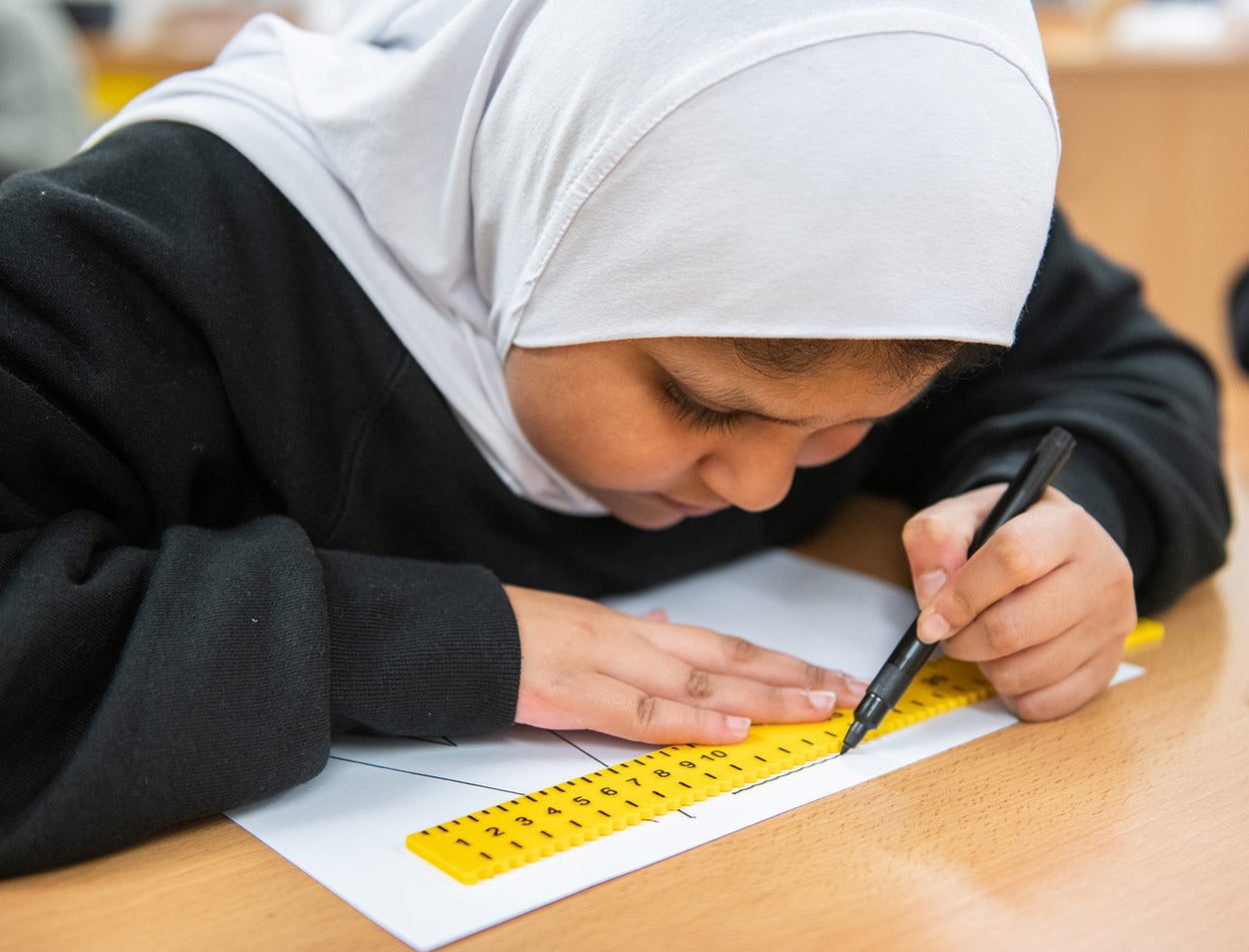 A pupil in black sweatshirt and white hijab uses the RNIB tactile ruler to draw a line