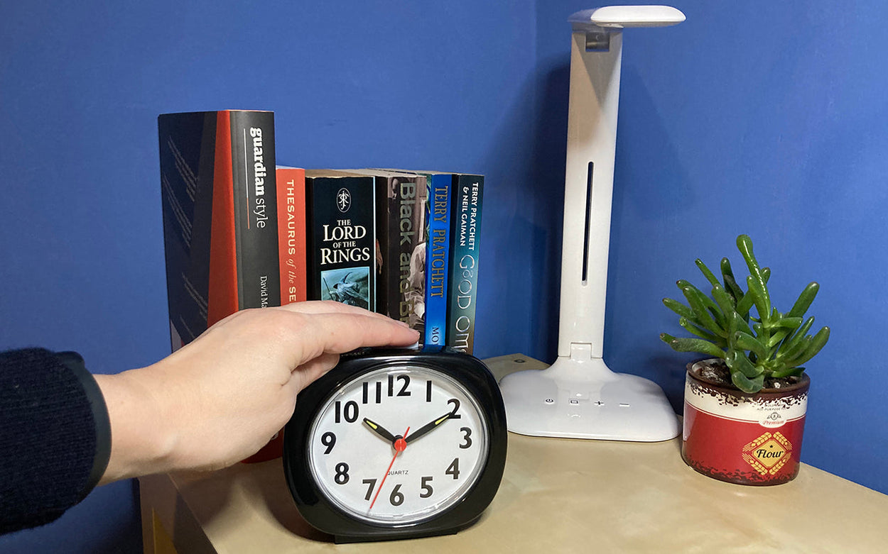 A person pressing the 'speak' button on top of RNIB's talking analogue clock which is on a bedside table with books and a lamp