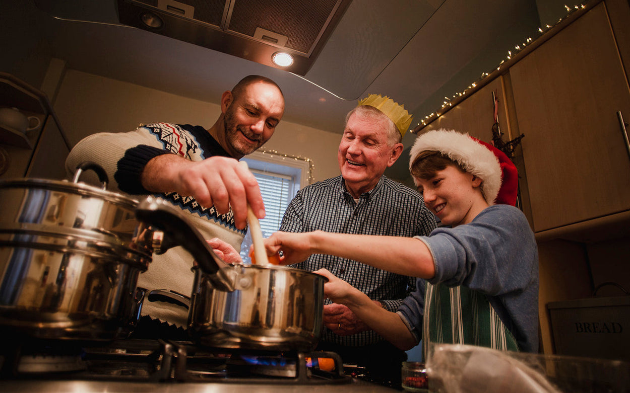 A middle-aged man in a Christmas jumper, an older man wearing a paper crown, and a teenage boy in a Santa hat are cooking at a gas hob