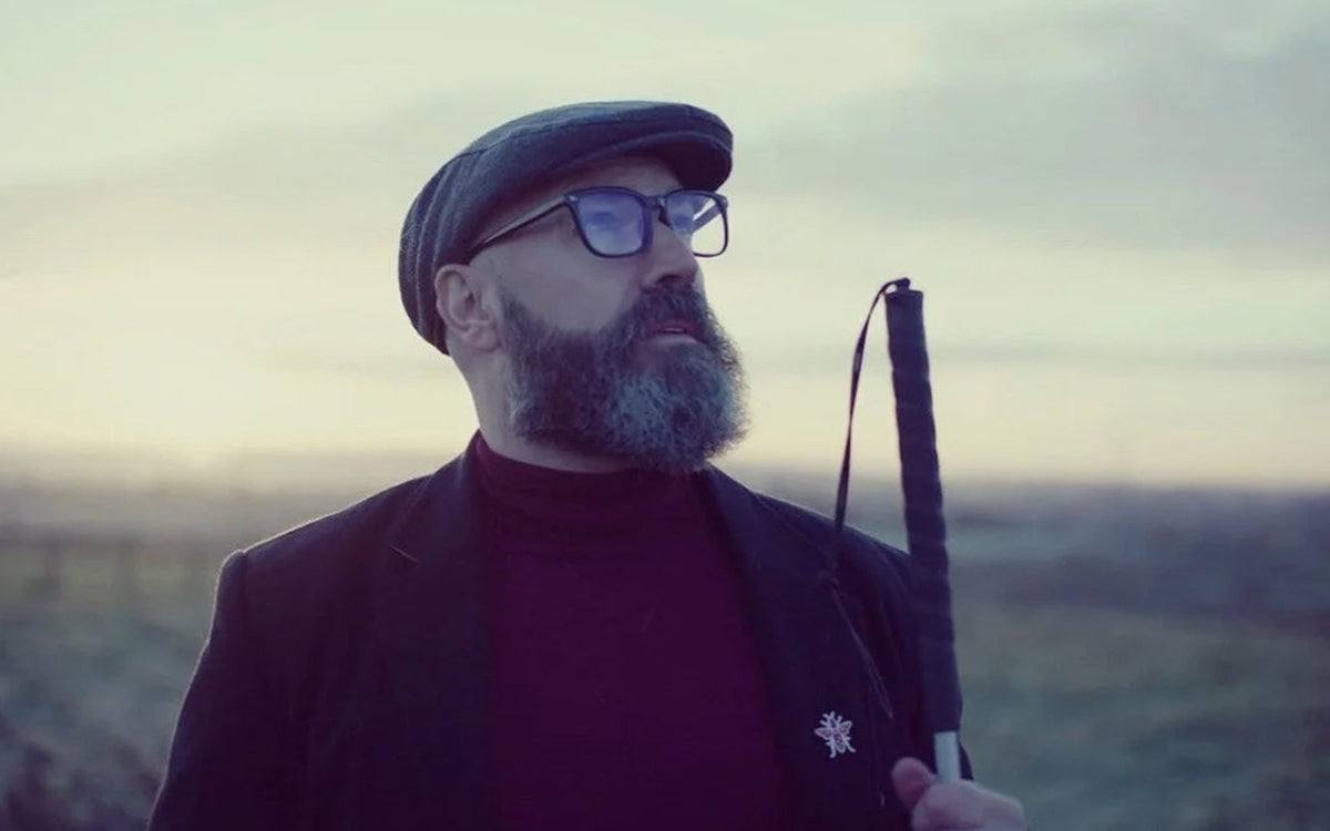 Dave Steele, the Blind Poet, in low light in front of a flat rural landscape that is out of focus. He's a white man with a bushy beard, wearing a burgundy rollneck, dark jacket, flat cap, and spectacles, who is holding a long cane.