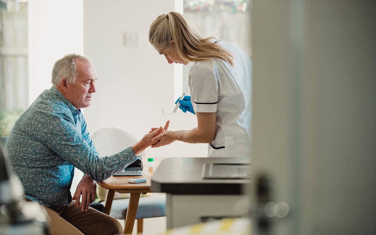 A nurse prepares to do a finger-prick test on a man sitting at a kitchen table.