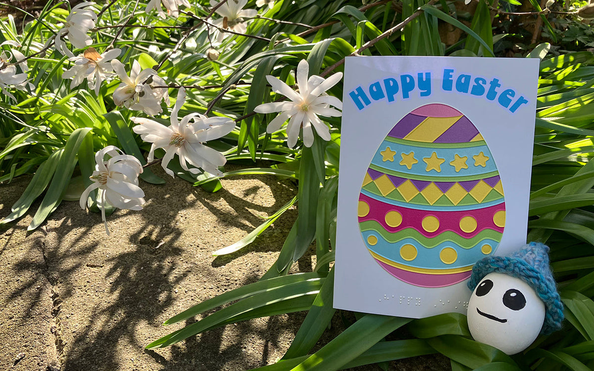 Colourful, tactile Easter egg card nestled in greenery with white magnolia blossoms. A white painted egg with big eyes and a small smile, wearing a knitted hat, sits near the card.