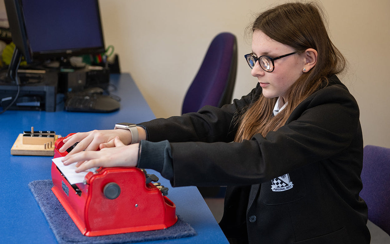 A girl in a black school blazer checks the braille she's produced on a red braille typewriter.