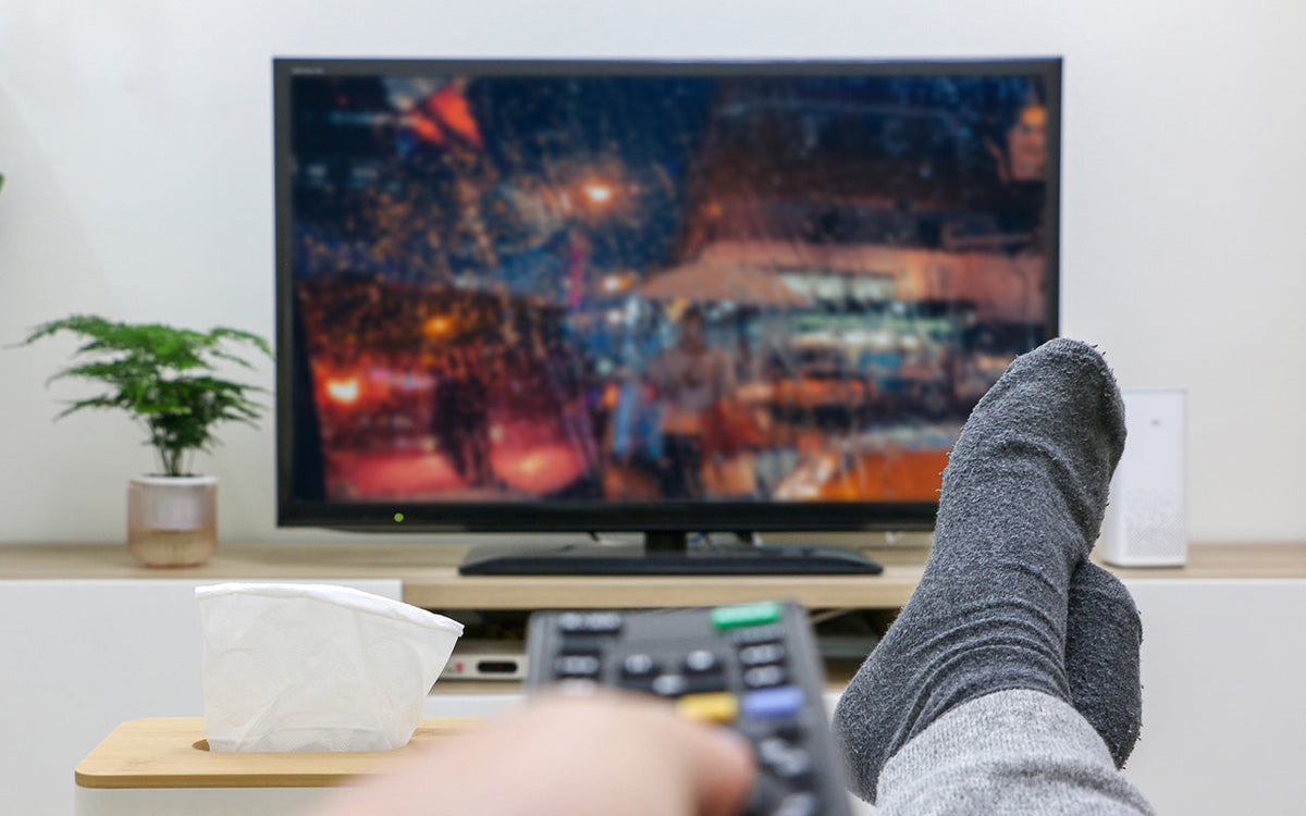 Image shows a person's point of view from a sofa looking at a TV screen. Their feet are up and they are pointing a remote control at the TV