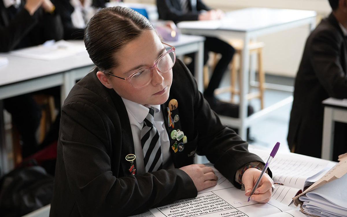 A girl in black school blazer listens attentively while working on a large print worksheet