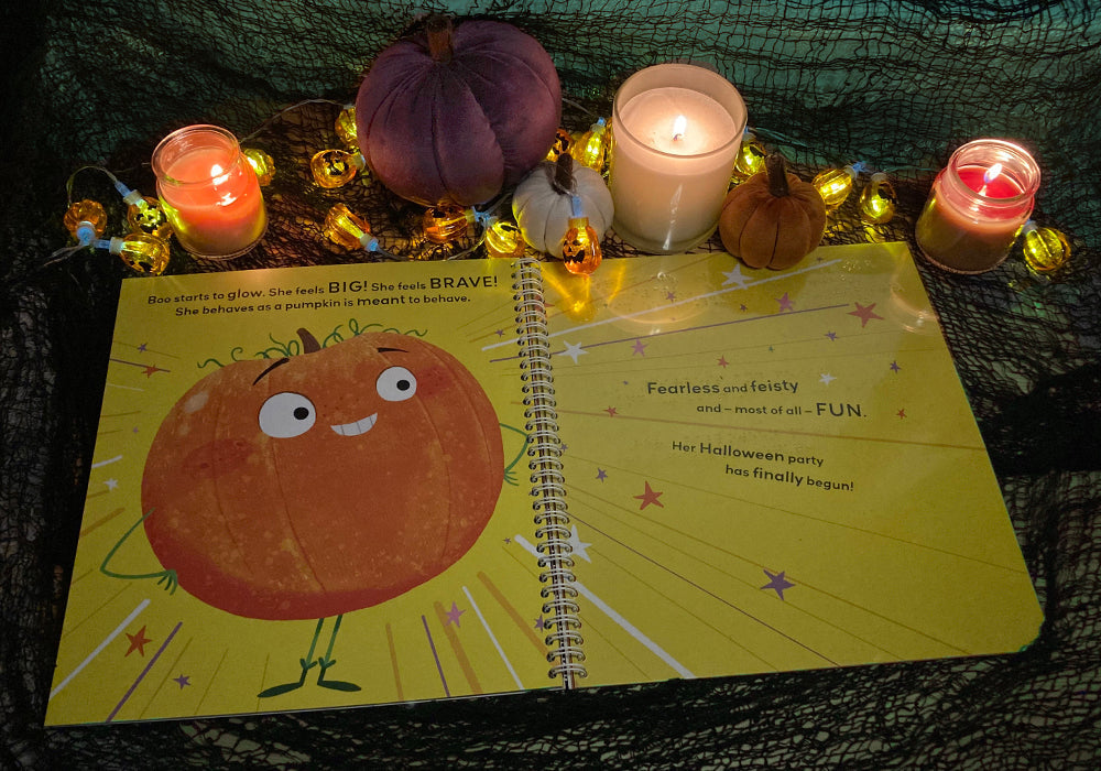 Braille and print picture book open to show a smiling pumpkin. The book is surrounded by plush pumpkins, candles and black web-like material