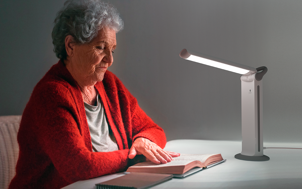 An older white woman in a red cardigan sits side-on to the camera reading a book on a desk using an adjustable Daylight Twist lamp for extra light