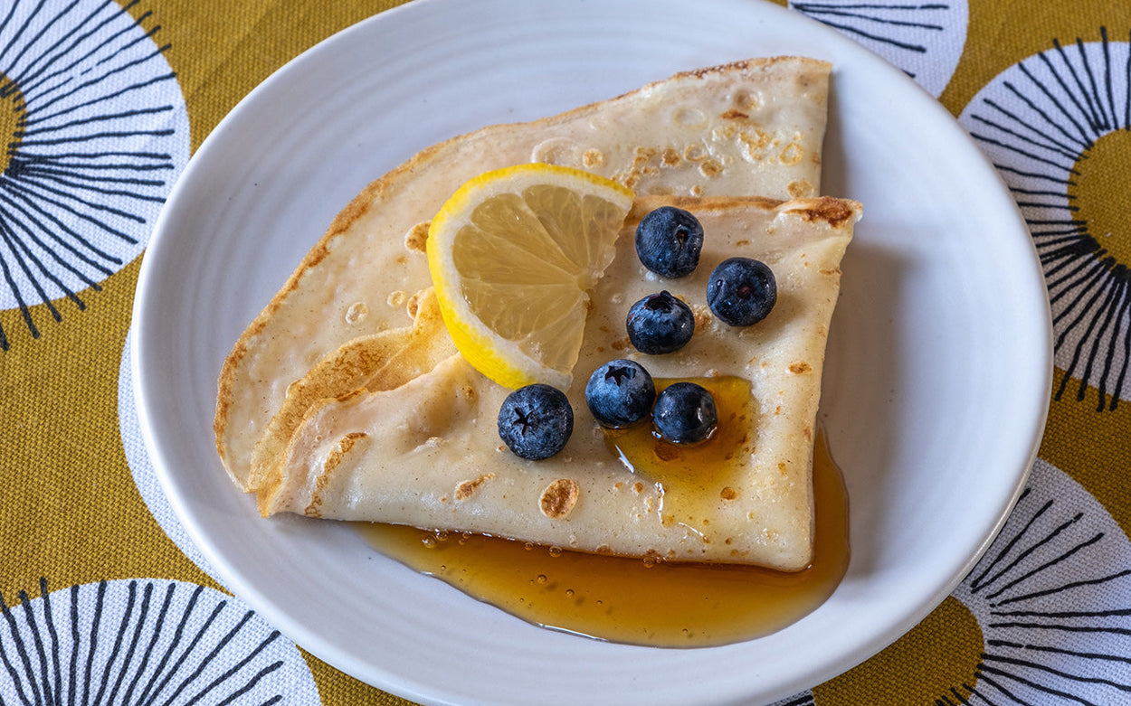 A pancake folded in quarters on a white plate and topped with syrup, blueberries and a wedge of lemon