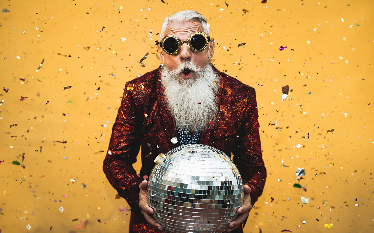 An older man with long, bushy white beard in sparkly gold-rimmed goggle-style sunglasses and red sequin jacket holds a glitterball while shiny confetti falls around him. He looks pleasantly surprised.