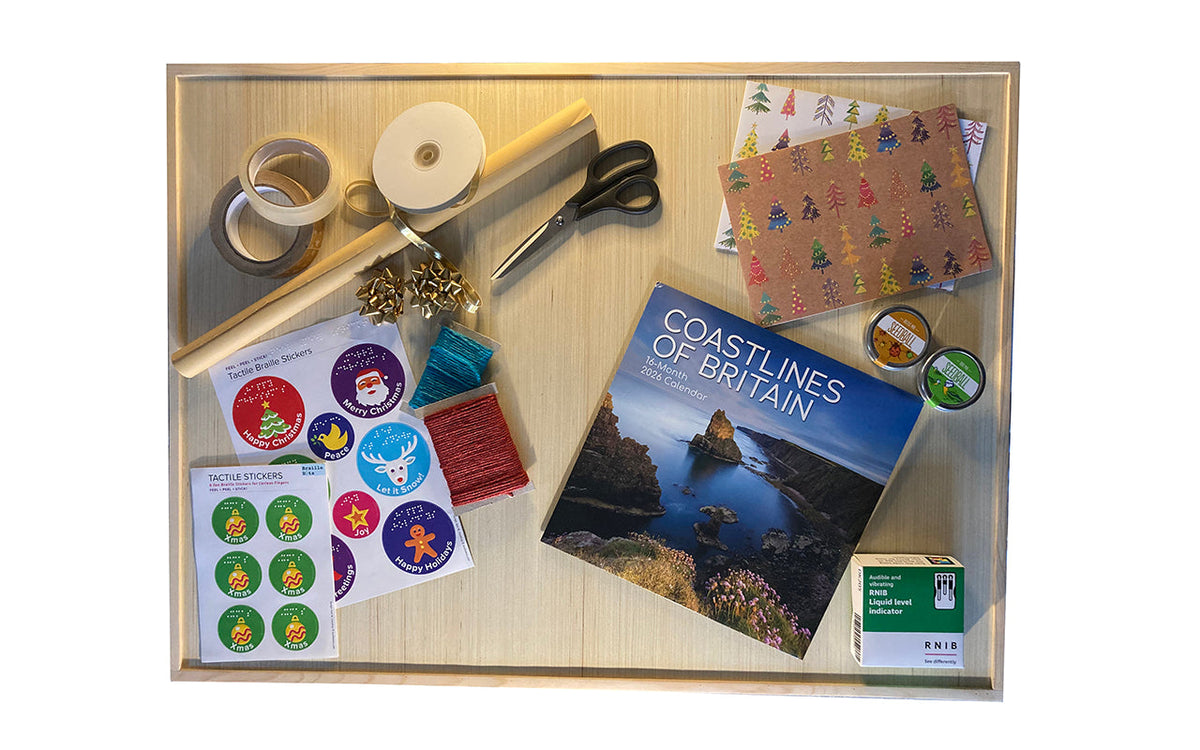 Gifts, gift wrap, tape, ribbon, coloured twine, braille gift stickers and scissors on a wooden board with a raised edge.
