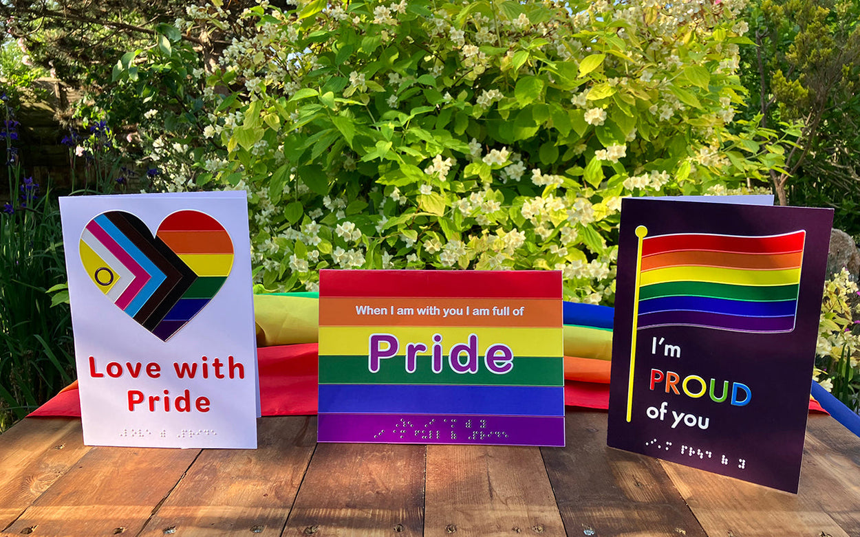 Three inclusive Pride cards with a draped Pride flag on a wooden table in a sunny garden.