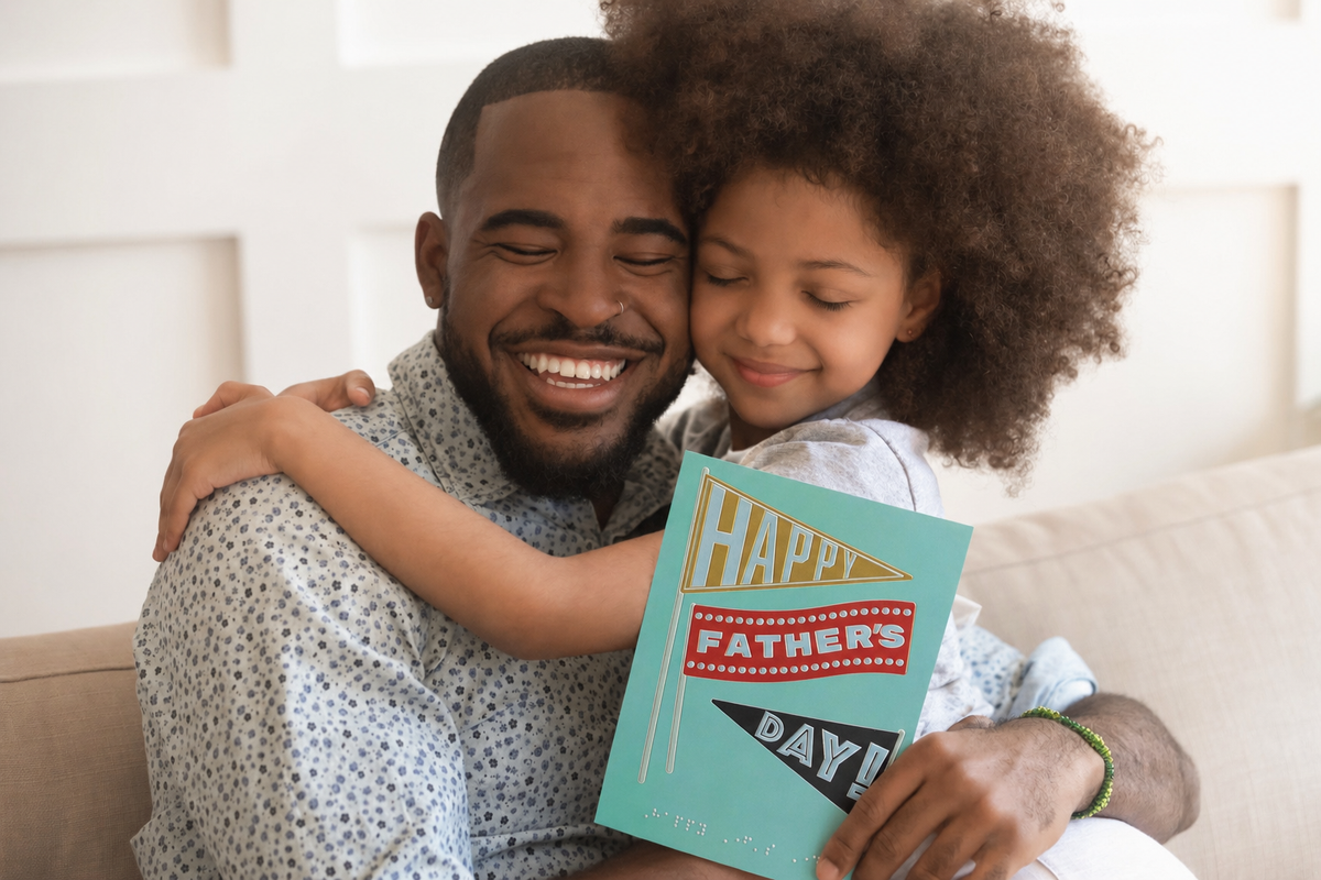 A father and daughter hugging and smiling with eyes closed. The father is holding RNIB's inclusive flag design Father's Day card in one hand.