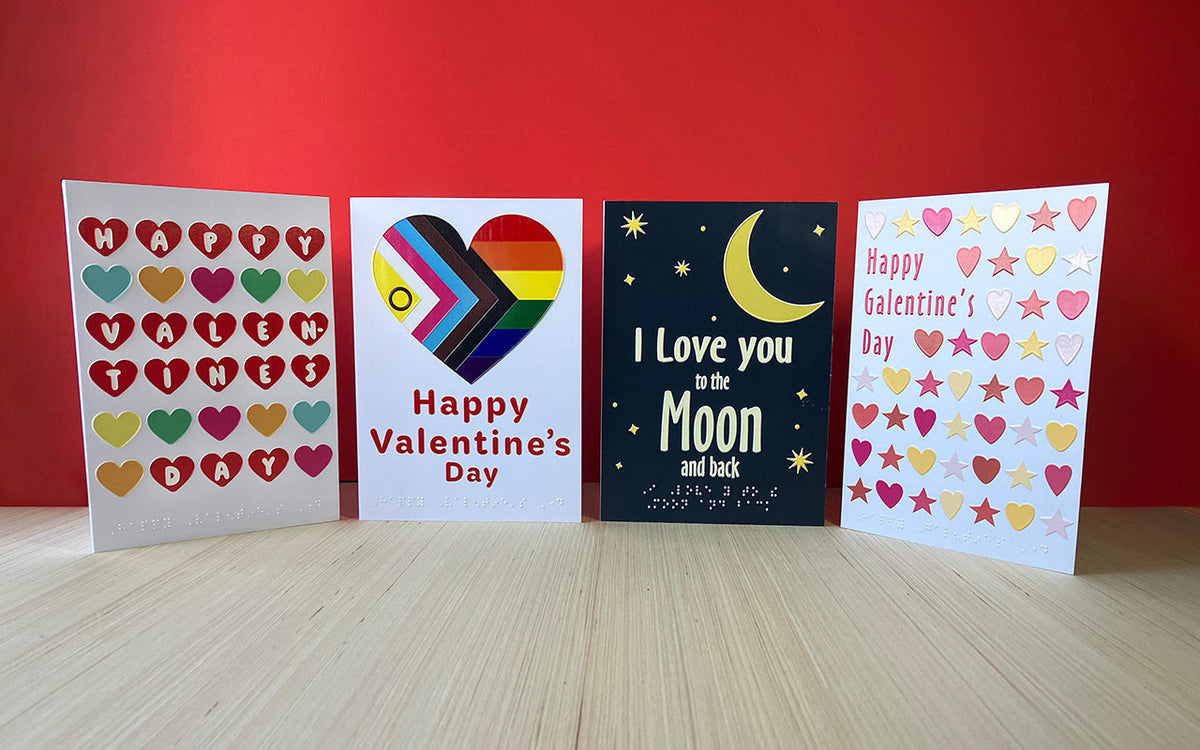 Four inclusive greetings card on a wooden surface in front of a red background. From left: A Love Hearts-inspired design, an intersex Pride Progress flag in a heart shape, a ‘I love you to the moon and back’ message, a Galentine’s Day card
