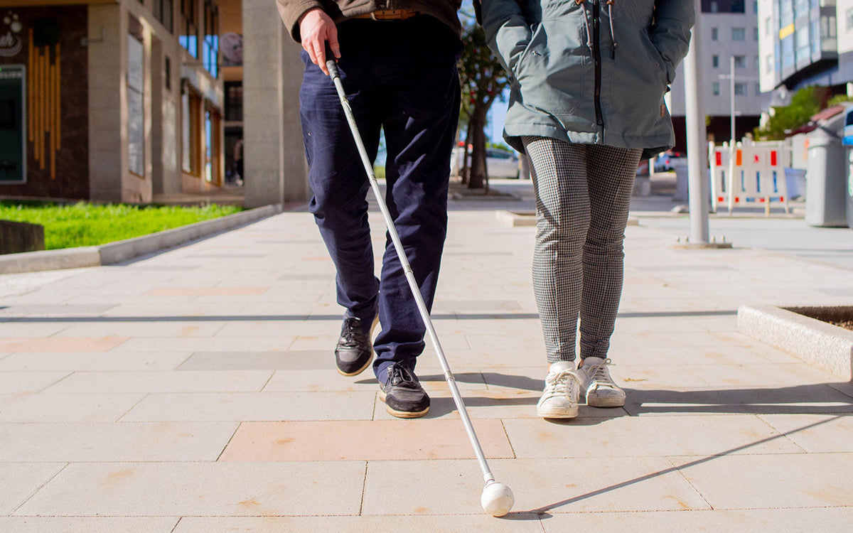 Two people walking on a paved surface towards the camera are visible from the waist down. One is using a long cane with a roller ball tip. The other is standing close by, likely acting as a guide.
