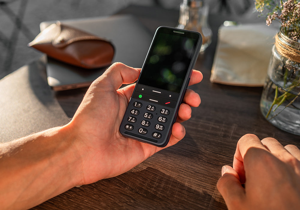 A hand holds a blue BlindShell Classic 3 mobile phone at a wooden table. Low sun illuminates the hand and a glass jar containing wildflowers.