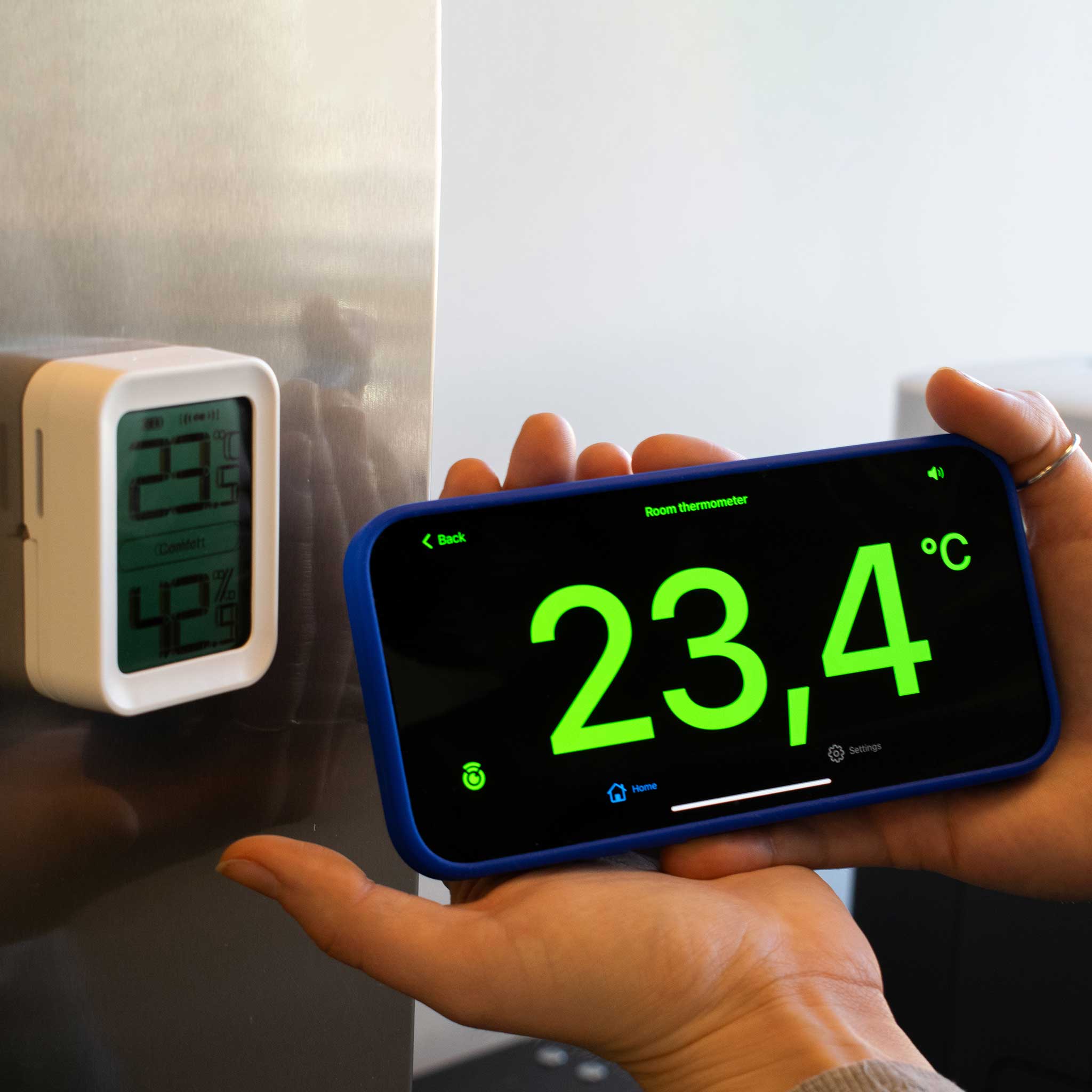Person holding a smartphone displaying temperature readings next to the Ikonn indoor thermometer on a fridge door.
