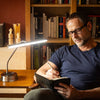 A man wearing black rectangular eyeglasses sits in a chair in front of a wooden bookshelf, writing in a black notebook with a pen. A modern silver and black desk lamp is turned on on a wooden table to his left.