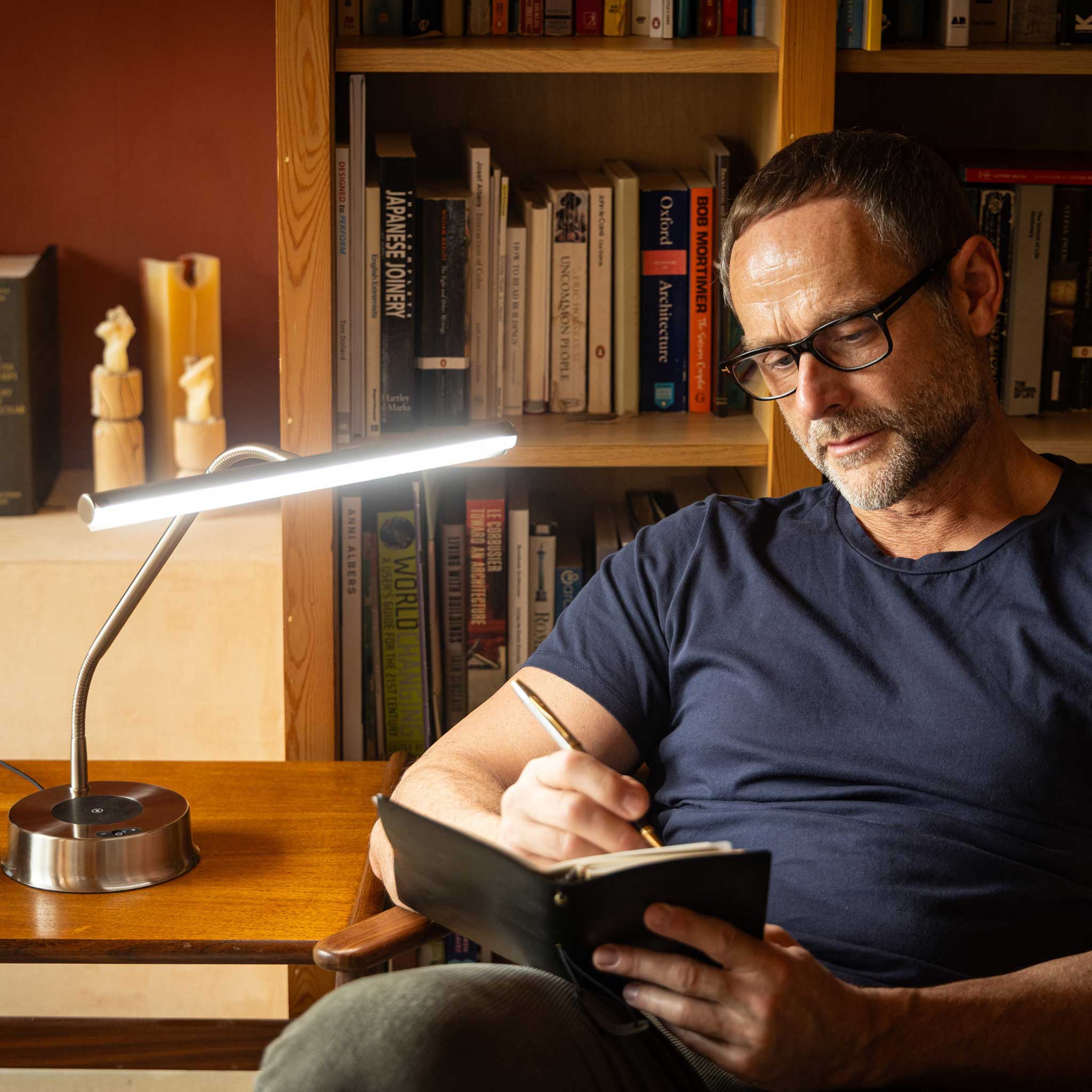 A man wearing black rectangular eyeglasses sits in a chair in front of a wooden bookshelf, writing in a black notebook with a pen. A modern silver and black desk lamp is turned on on a wooden table to his left.