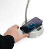 A silver wireless charging stand with a flexible gooseneck lamp and a smartphone placed on top, shown with a hand interacting with the base. The phone screen displays a green charging icon.