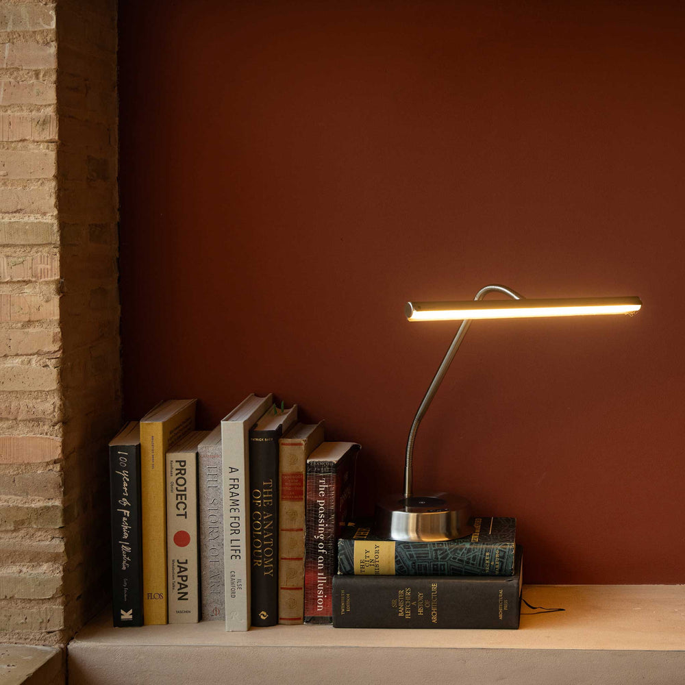 A modern LED desk lamp with a slim rectangular bar and curved metallic stem, glowing warm white on a shelf next to a stack of books against a dark reddish-brown wall.