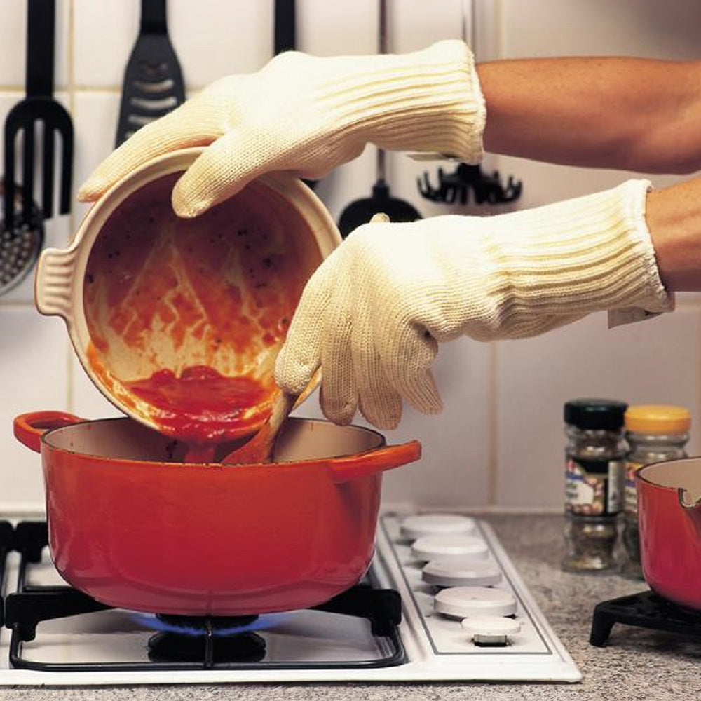 A person wearing the gauntlets to empty a small casserole dish into a larger one on a gas hob.