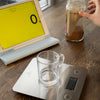 Glass mug on a digital kitchen scale next to a laptop with a yellow screen and a jar of coffee on a wooden surface.