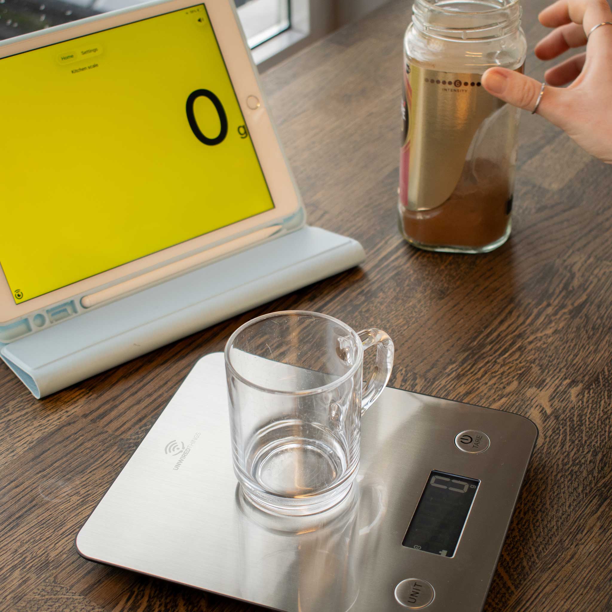 Glass mug on a digital kitchen scale next to a laptop with a yellow screen and a jar of coffee on a wooden surface.