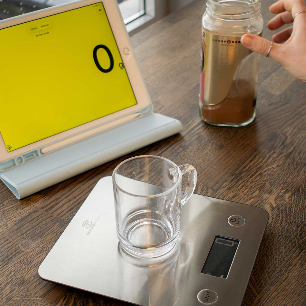 Glass mug on a digital kitchen scale next to a laptop with a yellow screen and a jar of coffee on a wooden surface.