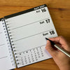 A spiral-bound weekly planner page from April 2027 is shown open on a wooden table, with a hand holding a pen pointing to Sunday 18th.