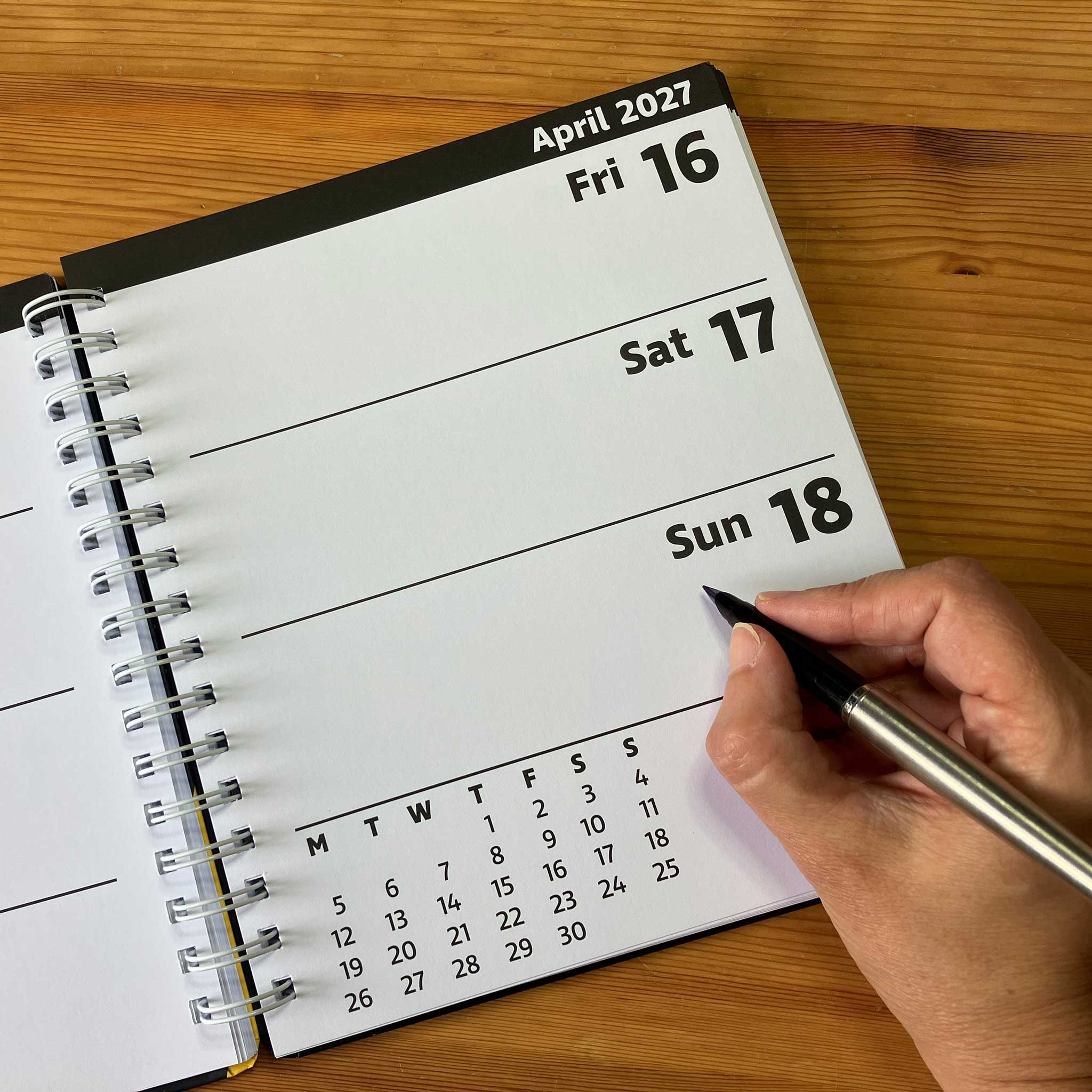 A spiral-bound weekly planner page from April 2027 is shown open on a wooden table, with a hand holding a pen pointing to Sunday 18th.