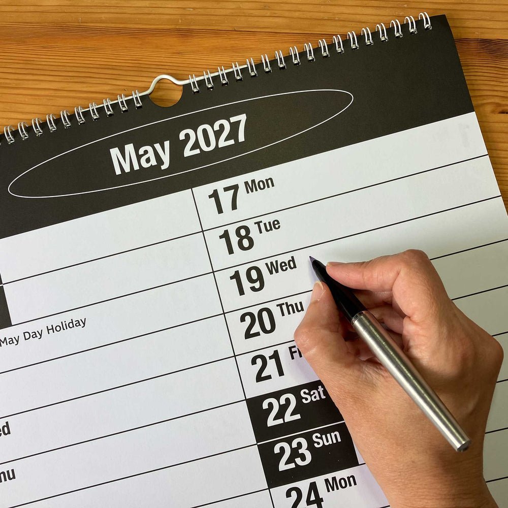 A spiral-bound May 2027 wall calendar page on a wooden surface, with a hand holding a silver pen marking the date 19 Wed.