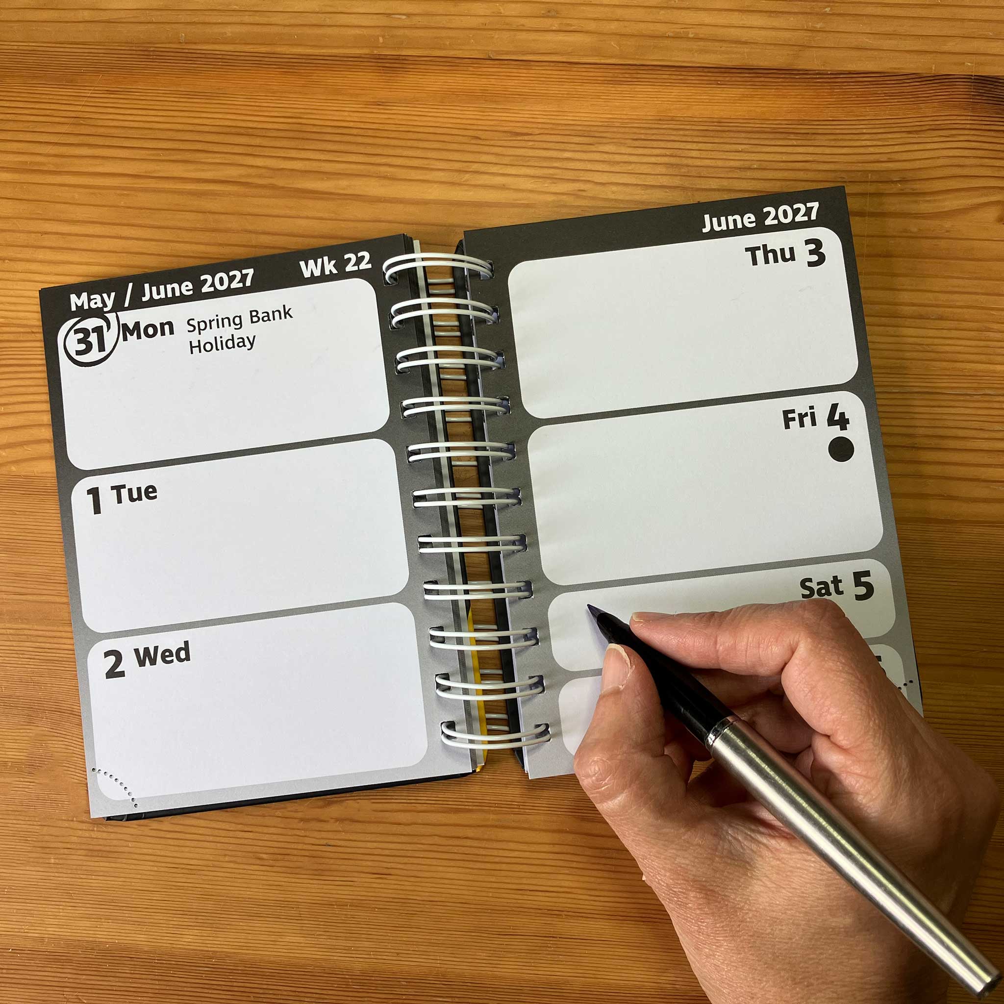 An open spiral-bound pocket diary on a wooden table, with a hand holding a black pen writing on one of the blank weekly boxes.