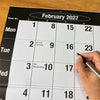 A close-up of a February 2027 wall calendar page on a wooden surface, with a hand holding a black pen pointing to the date February 23.