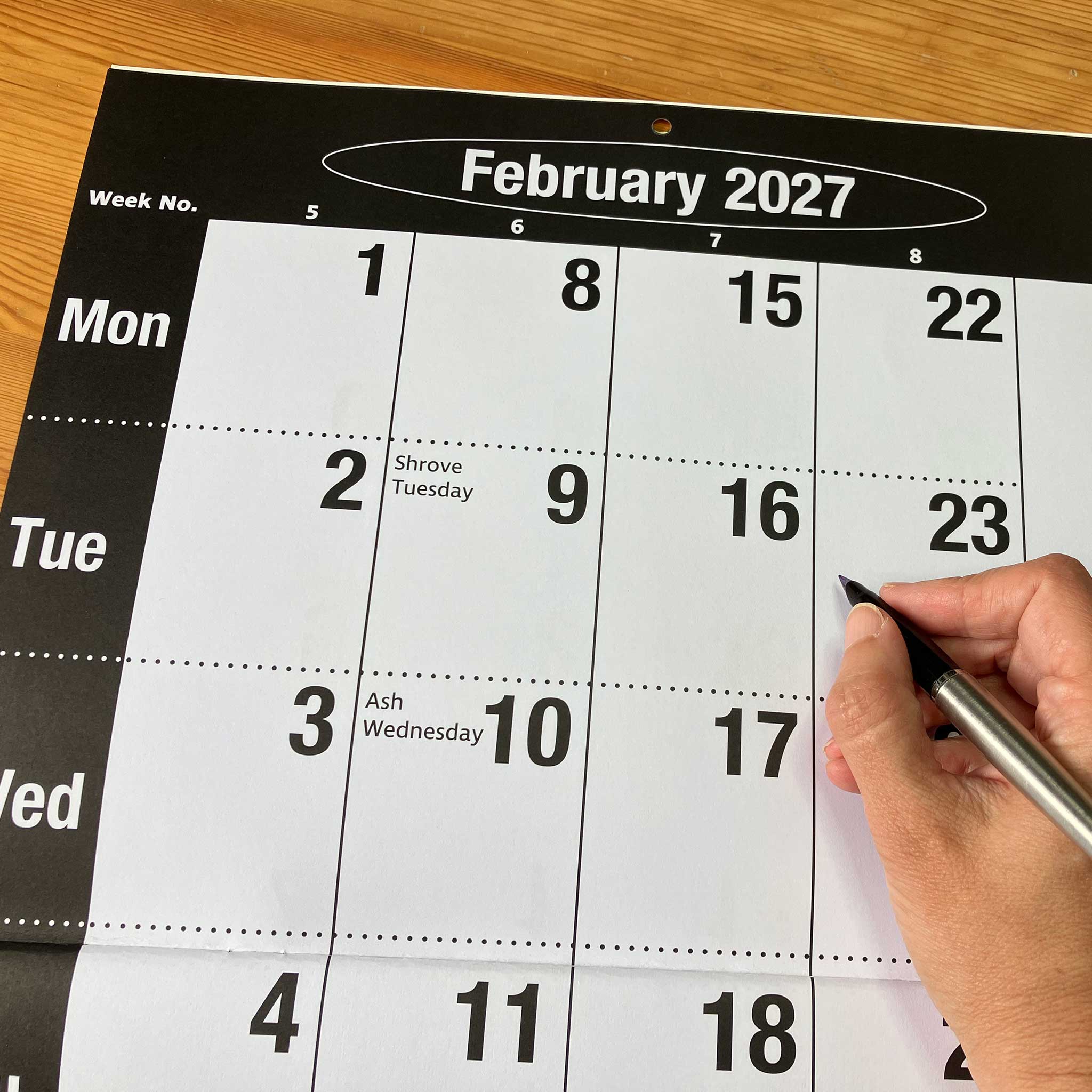 A close-up of a February 2027 wall calendar page on a wooden surface, with a hand holding a black pen pointing to the date February 23.
