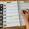 An open spiral-bound diary showing April with weekday columns and a notes page, with a hand holding a pen writing on the right side.