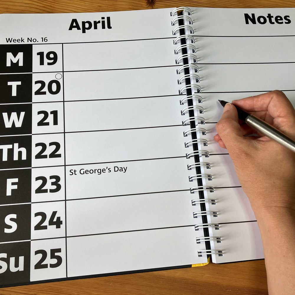 An open spiral-bound diary showing April with weekday columns and a notes page, with a hand holding a pen writing on the right side.
