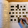 Crossword puzzle book with a partially completed crossword and a hand holding a pen.