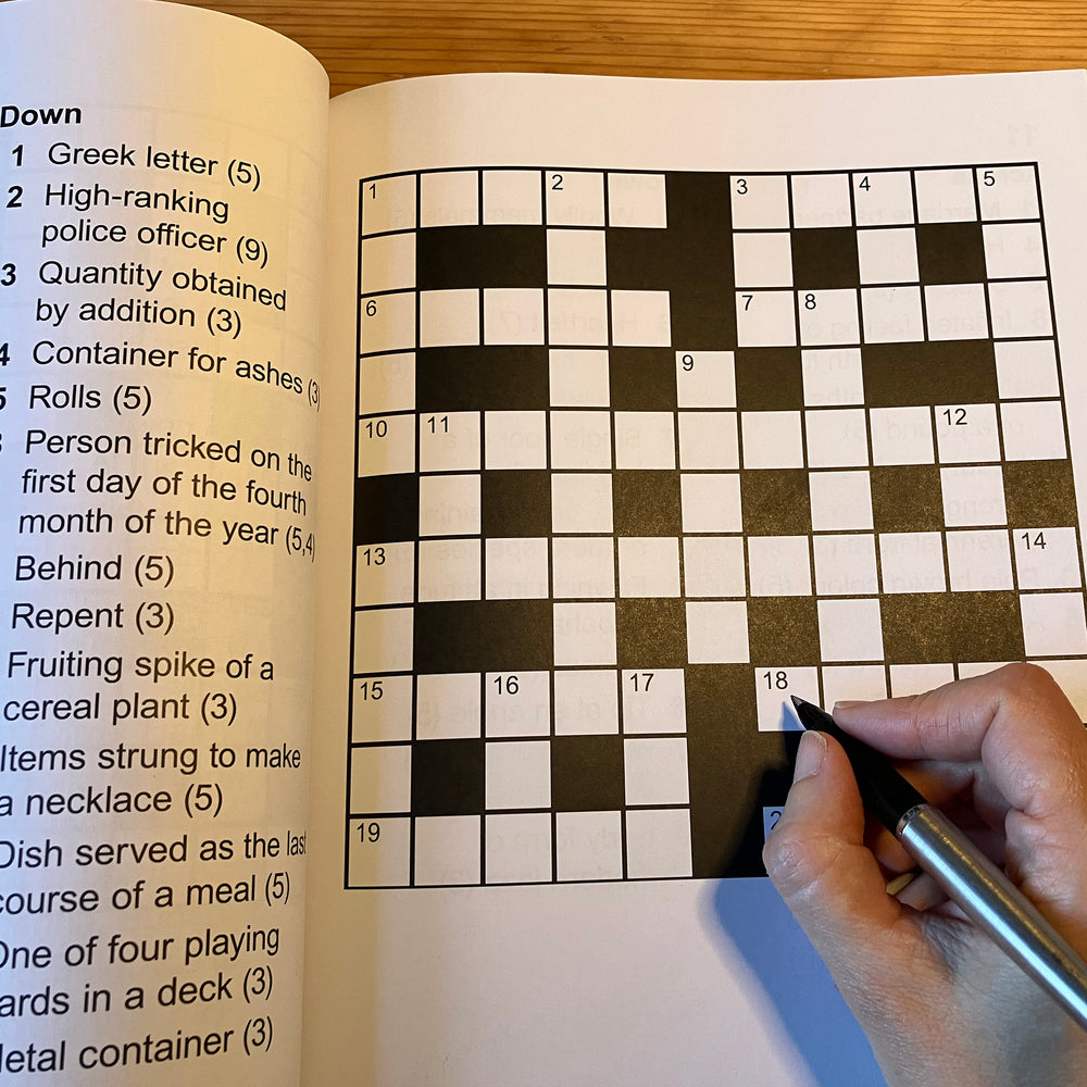 Crossword puzzle book with a partially completed crossword and a hand holding a pen.